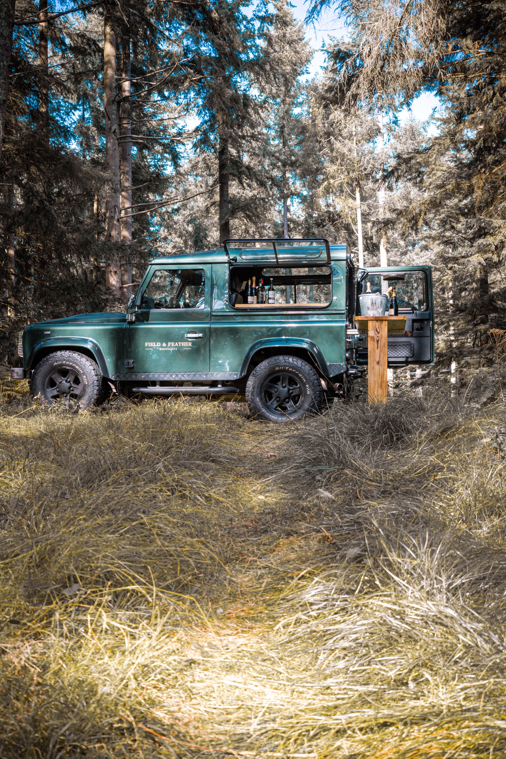 The Aintree Green Defender 90 in woodland with its bar open and oak serving table out